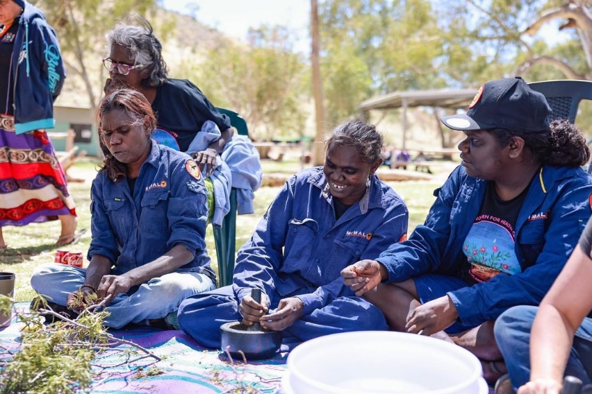 Three Indigenous rangers wearing vibrant blue shirts, sit on the ground and smile as they use traditional materials.