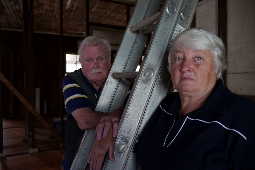 A man and a woman with white hair inside a gutted home next to a ladder