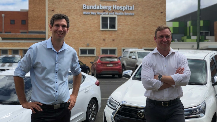 Two men wearing business shirts stand smiling towards the camera with the Bundaberg Hospital in the background.