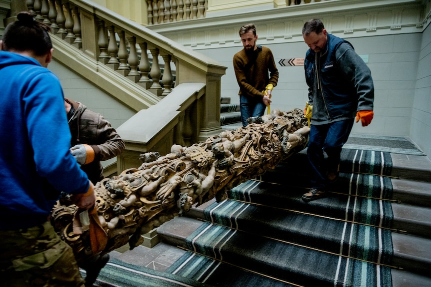 Four men wearing gloves carry a large cylindrical artwork down a set of carpeted stairs.