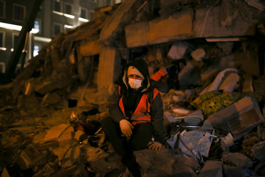 A person wearing high viz and a face mask sits on top of rubble. 