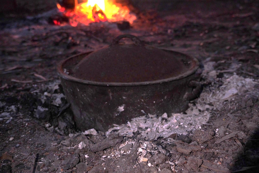 A pot with stew is in the ground, where hot ashes heat the dish. In the background a fire burns.