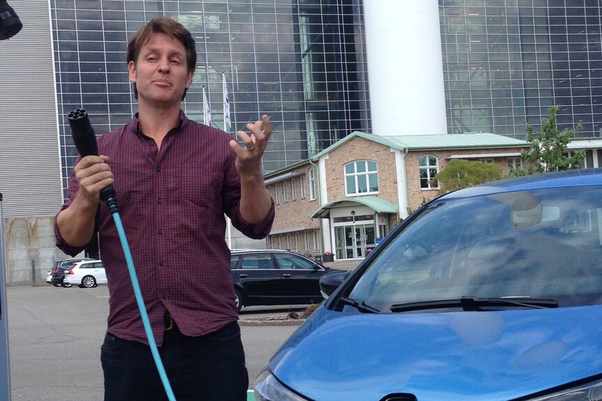 Craig Reucassel holds a large electric cable with one end plugged into a car, as he poses for the camera