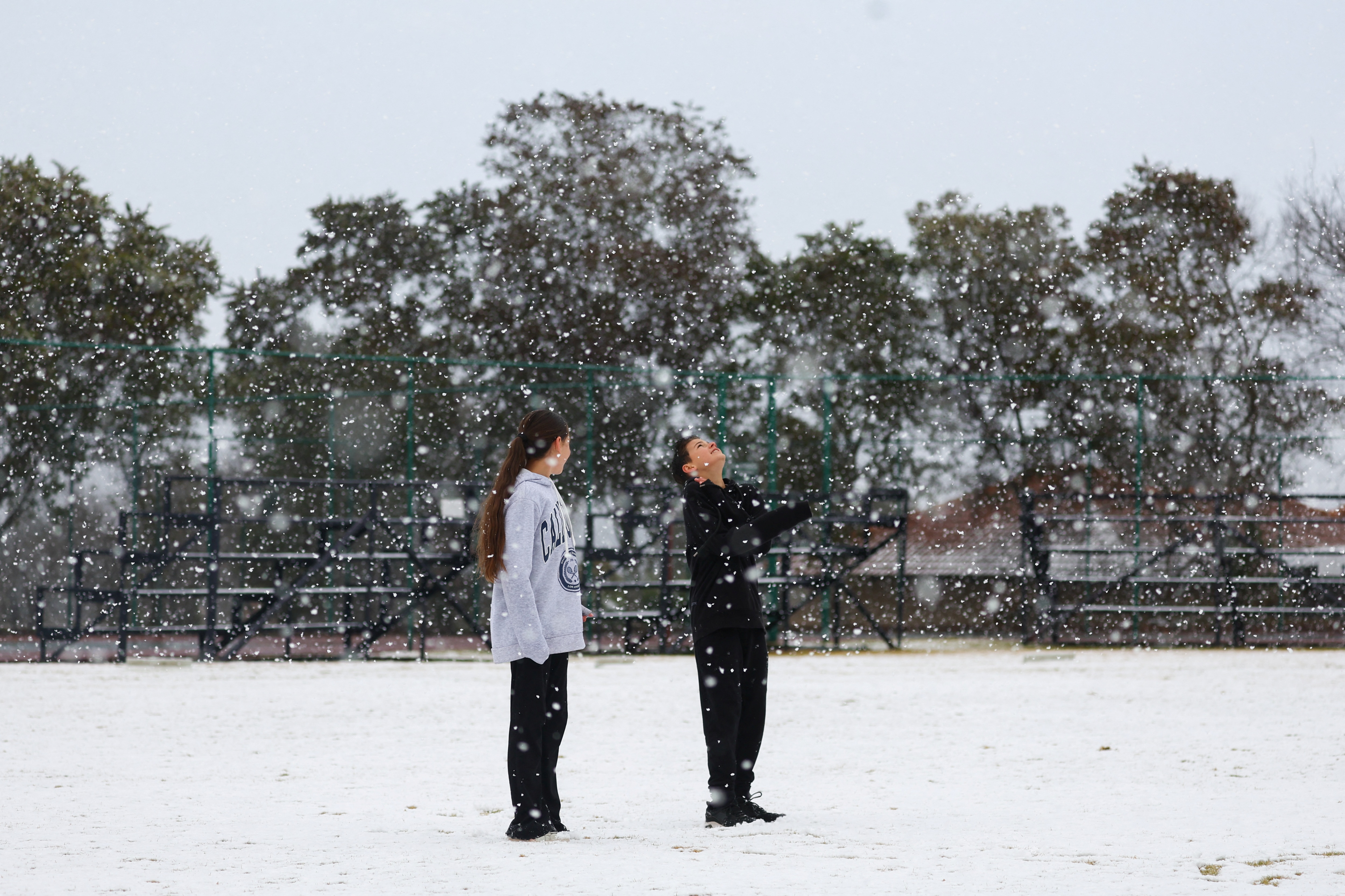 Two children stand side by side on snow covered ground, one child looks up at the falling snow. 