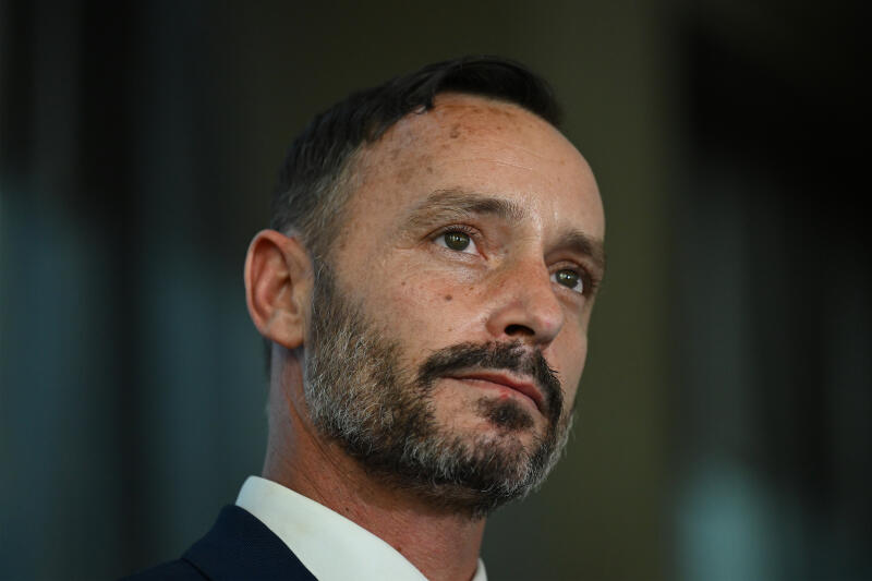 close-up image of a brunette man with dark facial hair looking into the distance
