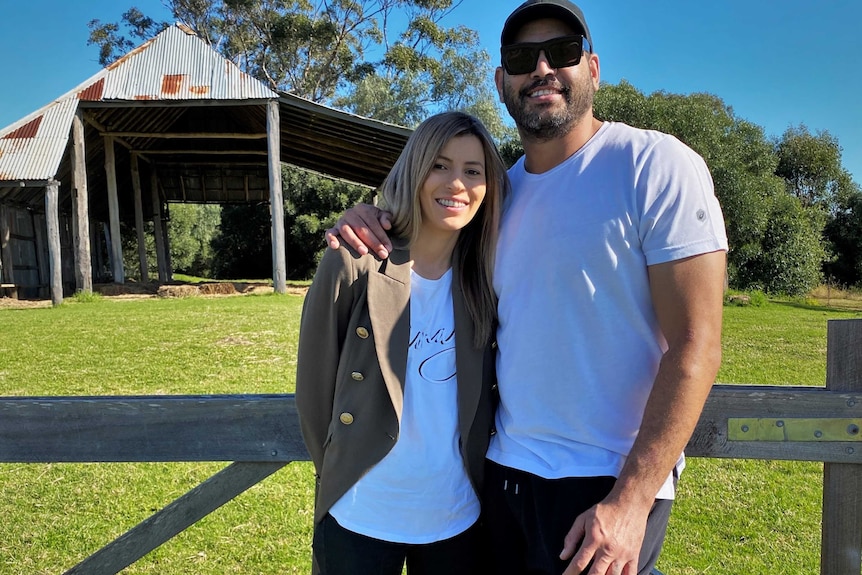 A man wearing a white t-shirt and sunglasses stands next to and has arm around woman in black jeans and brown jacket, smiling