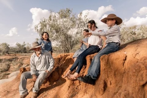 Portrait: Mum and Dad sit on the lower end of an embankment looking at their three kids slightly above them in the NT outback.