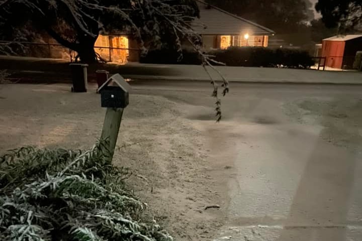 Snow across a road and driveway with house lights on in the background.