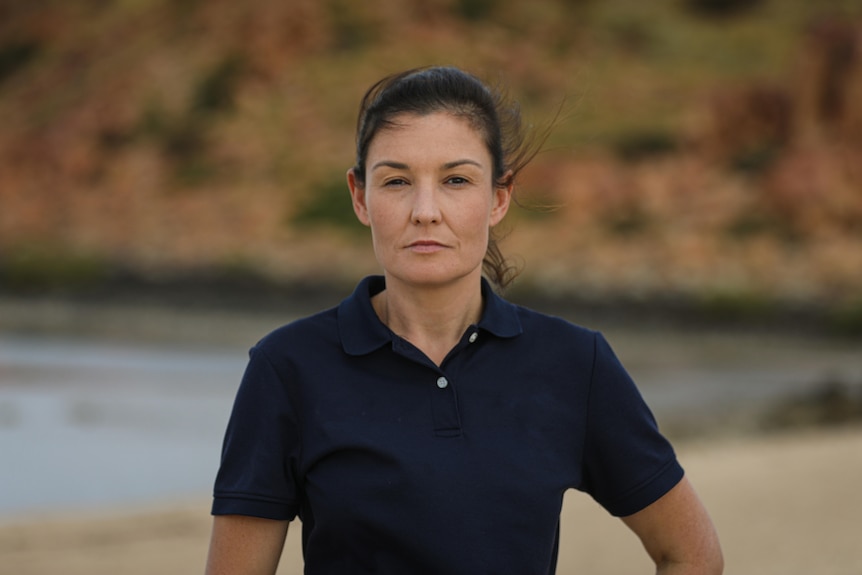 A serious woman, hair tied back, black T-shirt, with company logo, faces the camera, beach, rocky red outcrops in the back.