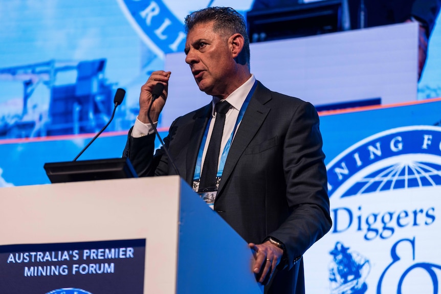 Un hombre vestido con traje y corbata hablando detrás de un podio en una conferencia minera.   