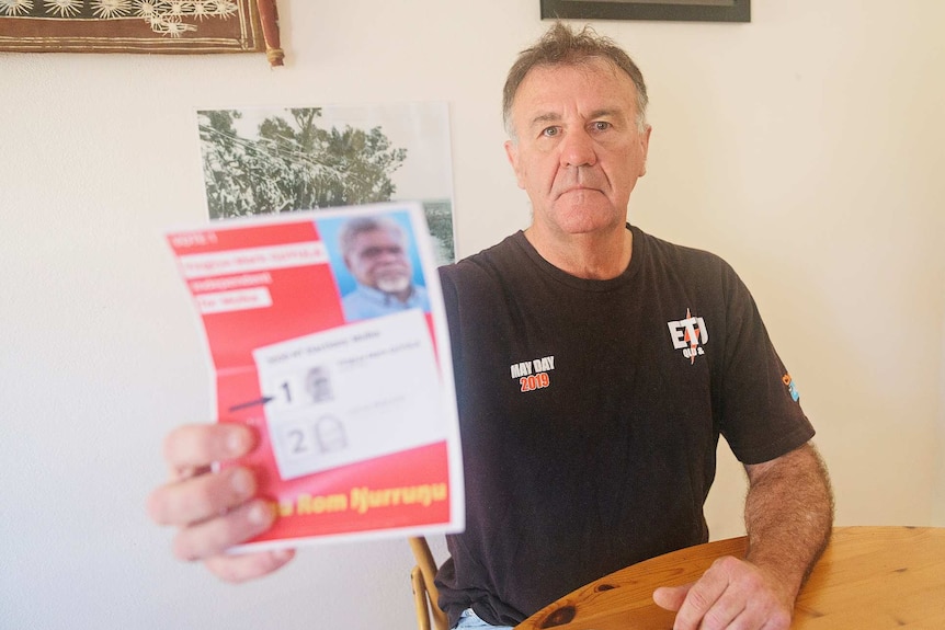 Dave Mitchell sits in a black top in a home holding up a red flyer with Yingiya Guyula printed on it.