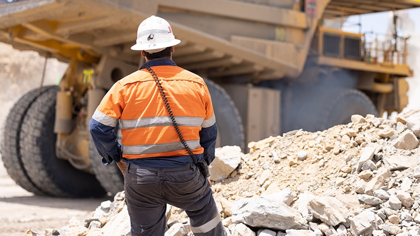 A man in an orange high-vis uniform and helmet stands in front of a large truck and pile of rocks