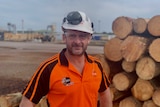 A man wearing high-vis and a hard hat stands in front of logs with a mill in the background.