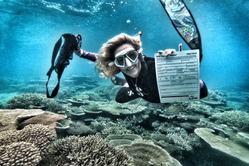 A woman scuba diving underwater smiles for the camera and shows off a certificate with coral below her.