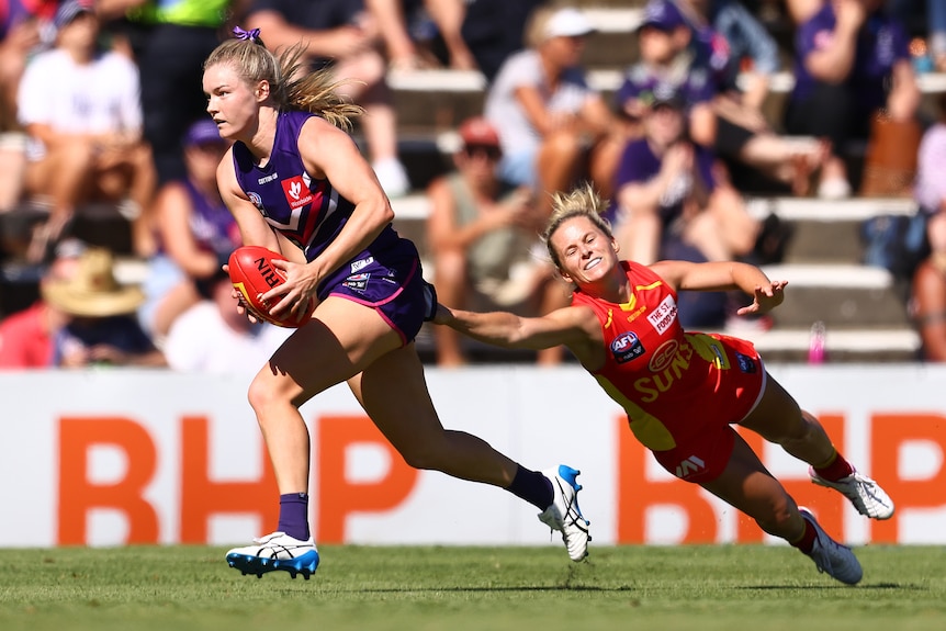 An AFLW player looks downfield running with the ball, as a defender throws out an arm while diving full-length to stop her.