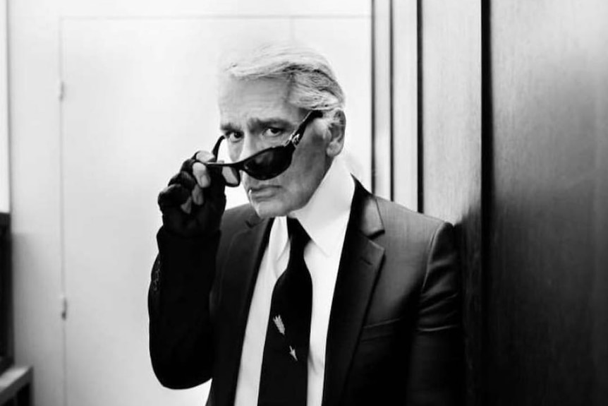 Black and white photo showing Karl Lagerfeld in a black suit and white shirt taking off sunglasses