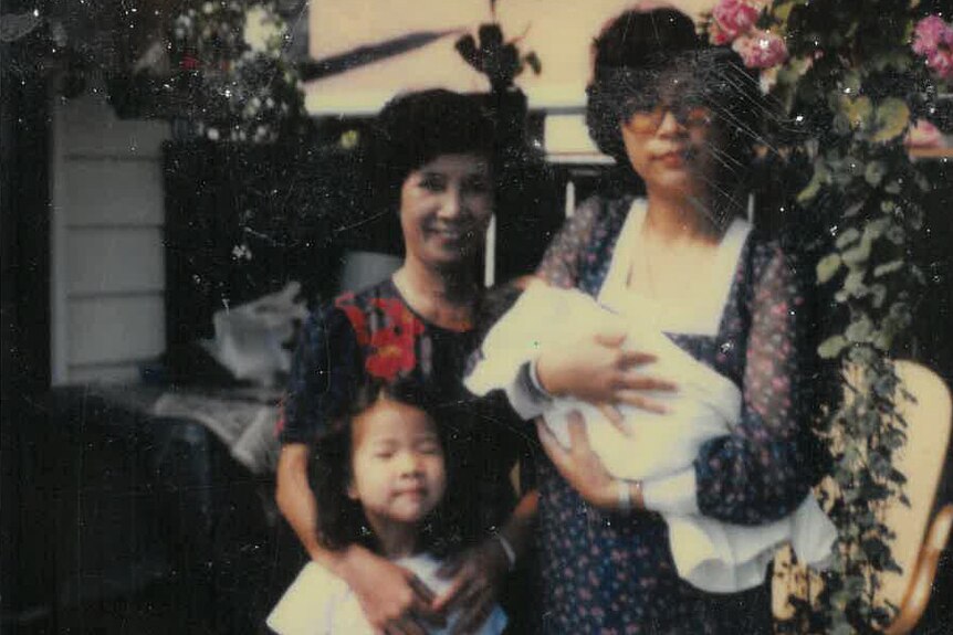 Slightly dark and grainy photo of Beverley Wang as young child standing with her mother holding a baby, and her smiling grandma.