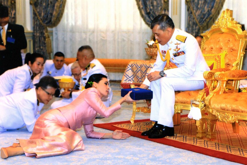 Thailand's new queen is seen bowing in front of the king who is blessing her while sitting on a chair.