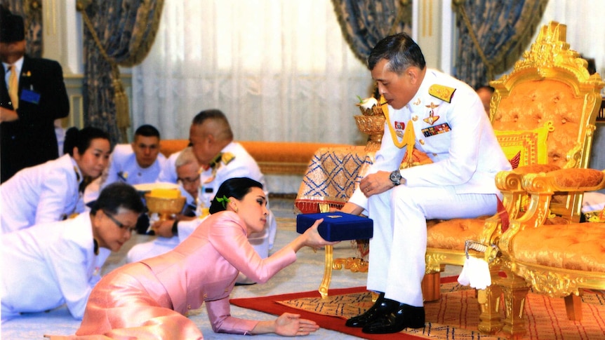 Thailand's new queen is seen bowing in front of the king who is blessing her while sitting on a chair.