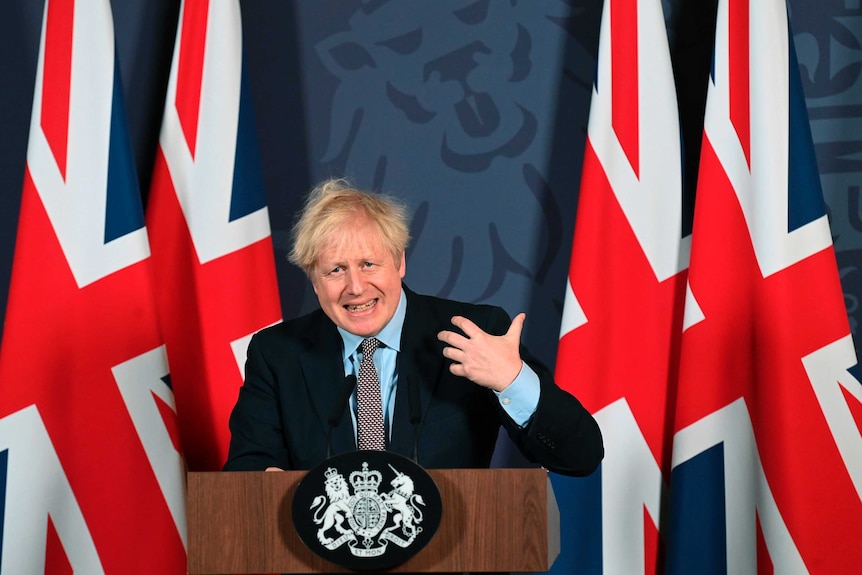 A man with blonde hair wearing a dark suit stands at a podium with British flags behind him.