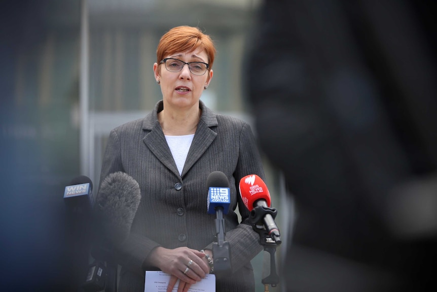 Rachel looks serious while addressing the media in a grey pantsuit.