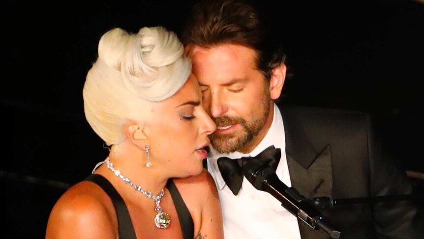 Lady Gaga and Bradley Cooper sing into a microphone seated next to each other