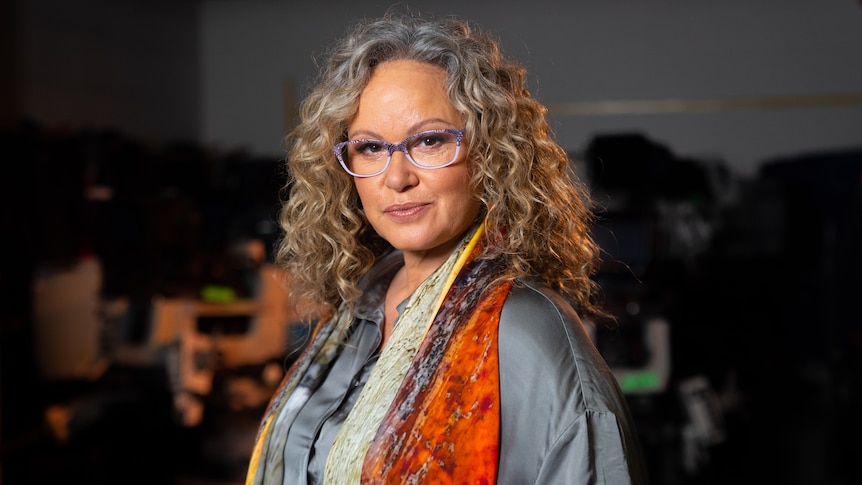 Portrait of Leah Purcell with purple thick rimmed glasses and burnt orange shawl looking straight with slight smirk on face