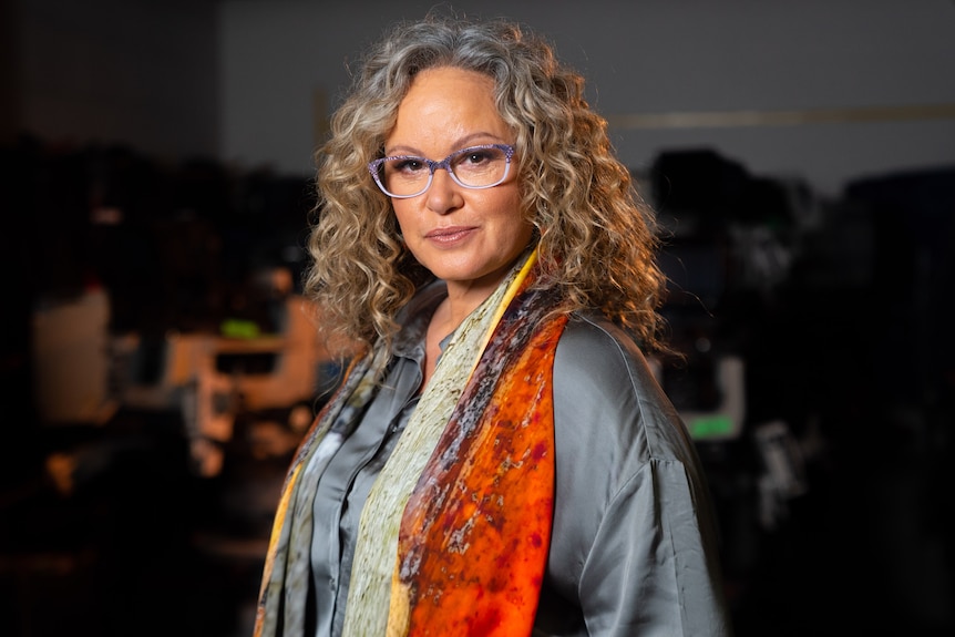 Portrait of Leah Purcell with purple thick rimmed glasses and burnt orange shawl looking straight with slight smirk on face