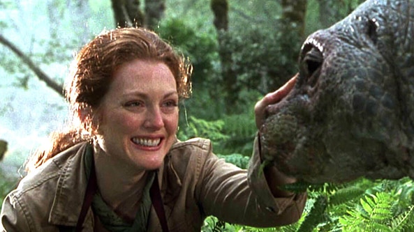 A woman with long red hair holds camera, smiles and pats dinosaur like creature in jungle on bright day.