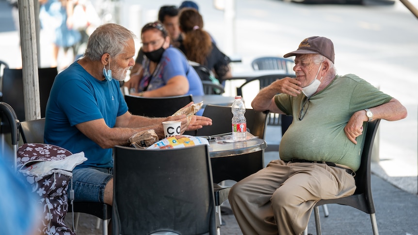 Two older men sitting at an outdoor cafe table with takeaway food. Both are wearing masks pulled down to their chins