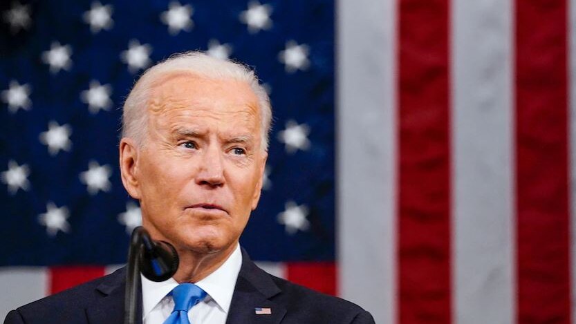 Joe Biden speaking into a microphone in front of an American flag