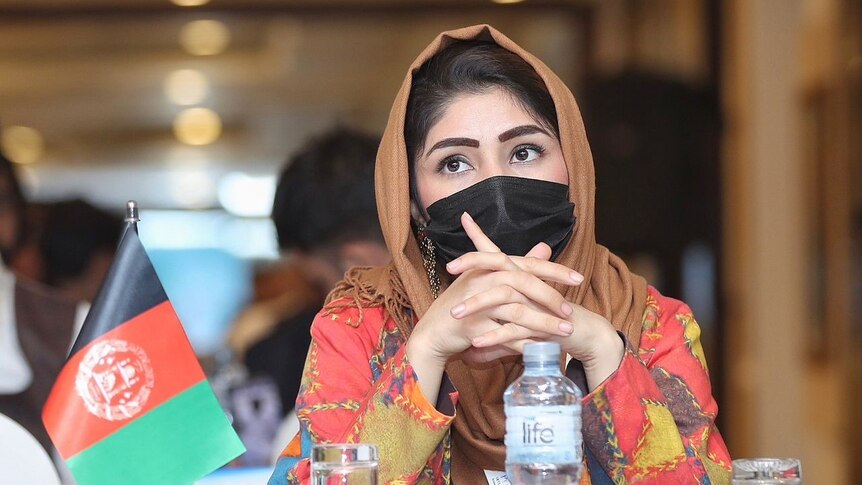 A young woman wearing a face mask and head scarf sits at a table with an Afghan flag