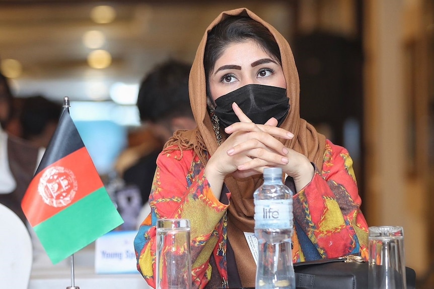 A young woman wearing a face mask and head scarf sits at a table with an Afghan flag
