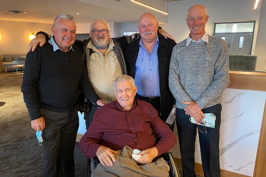 Four men standing behind older man sitting in wheelchair