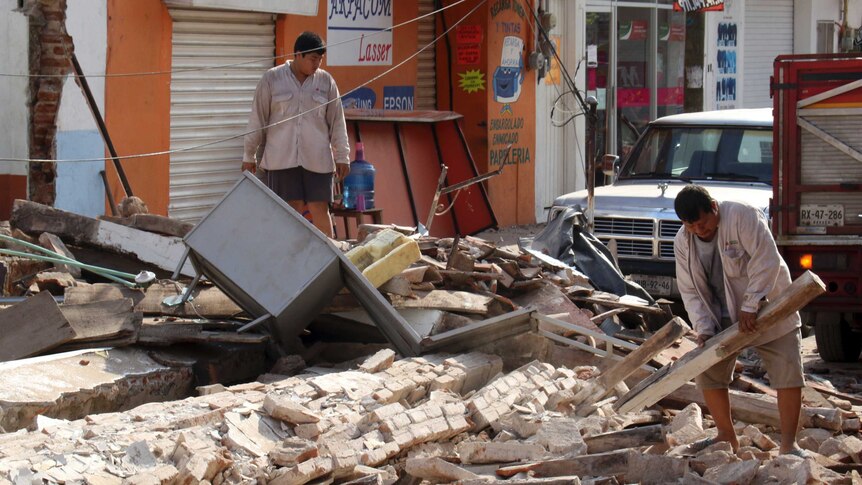 Two men clean up rubble from the front of a shop.
