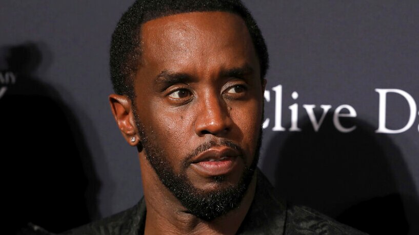 Sean Combs, wearing a black jacket, looks neutrally off camera in front of plastered logos.