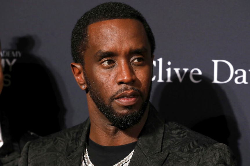 Sean Combs, wearing a black jacket, looks neutrally off camera in front of plastered logos.