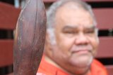 A man in the background looks ahead to the the camera past a traditional weapon called a Nulla Nulla