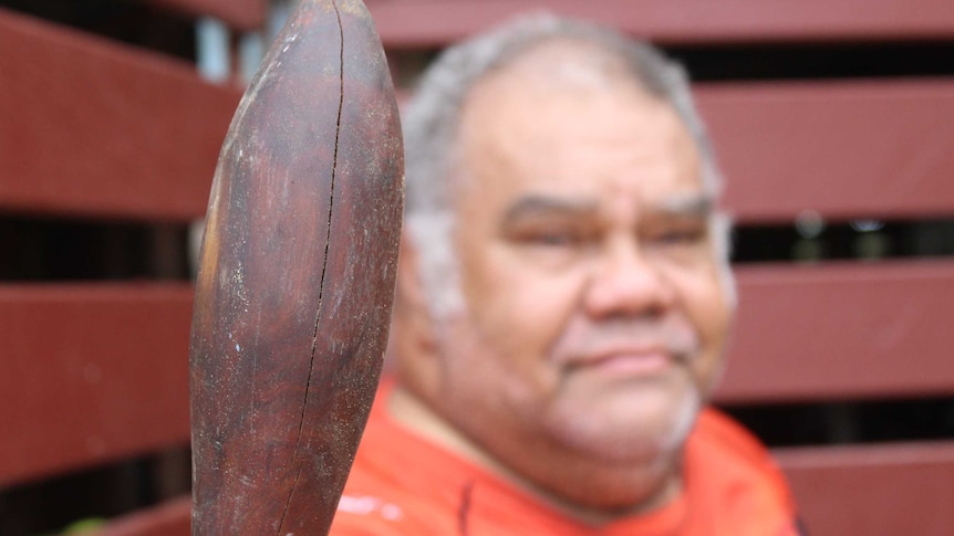 A man in the background looks ahead to the the camera past a traditional weapon called a Nulla Nulla