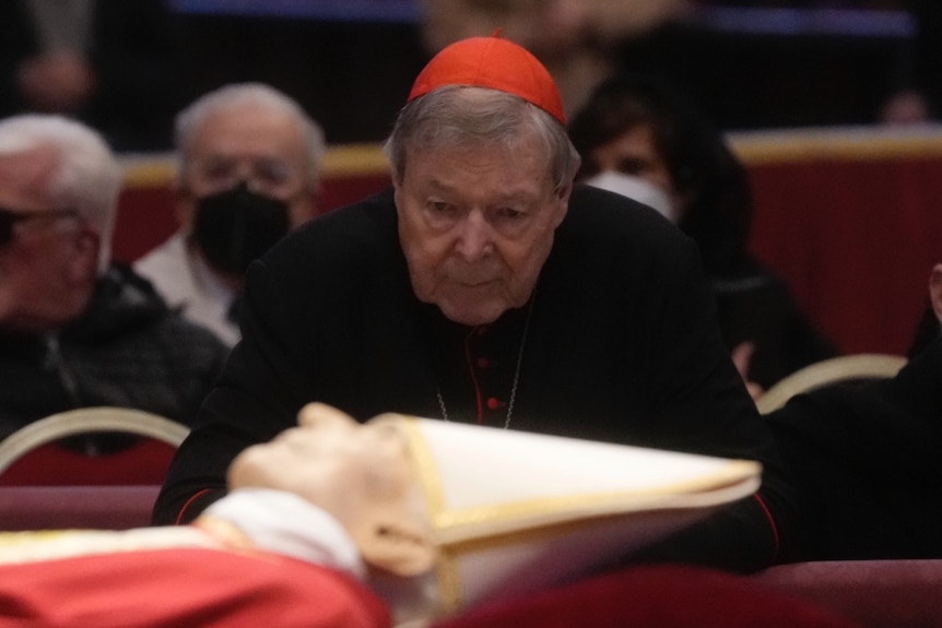 Pell, in a red cap, looks over the embalmed body of the former pope