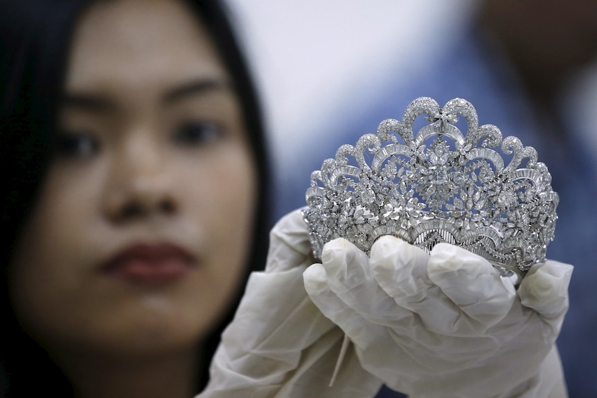 A young woman in white gloves holds a diamond tiara up to the camera