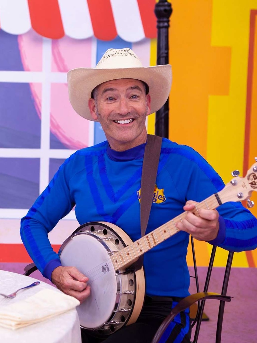 A man in a cowboy hat and blue skivvy plays a banjo and grins