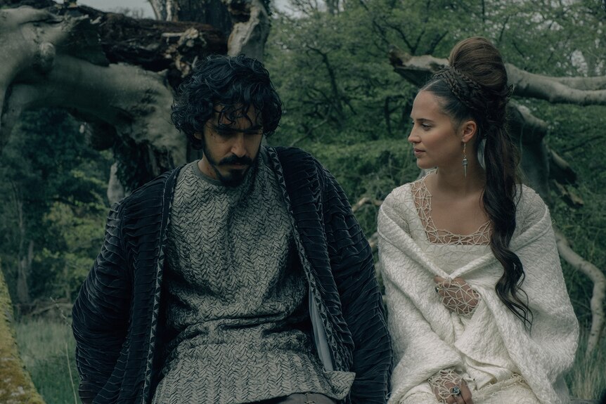 Young man with dark hair and beard and woman with long plaited dark hair are dressed in medieval garb in a wooded green forest.