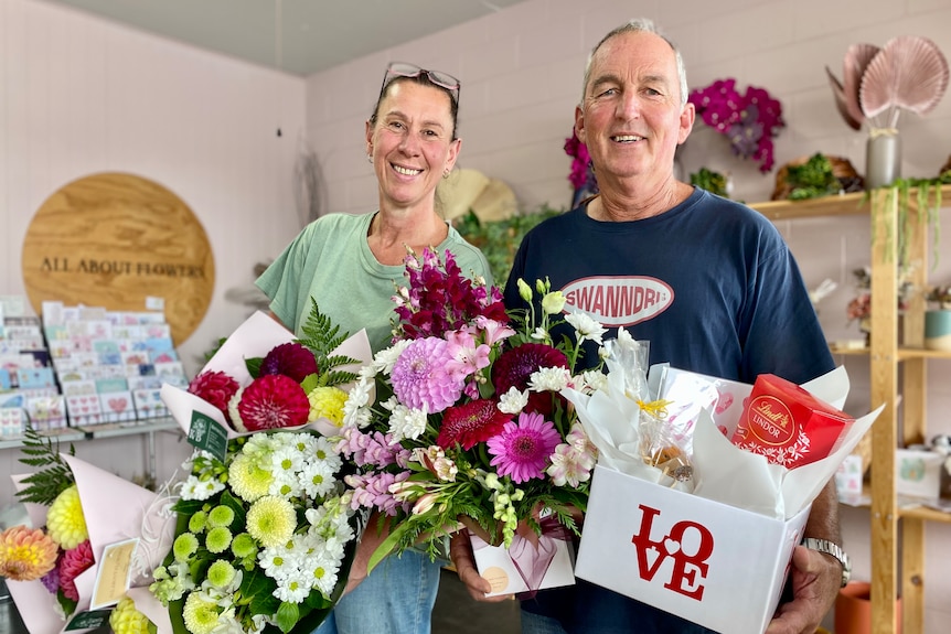 Two people stand inside a florist holding two large bunches of flowers