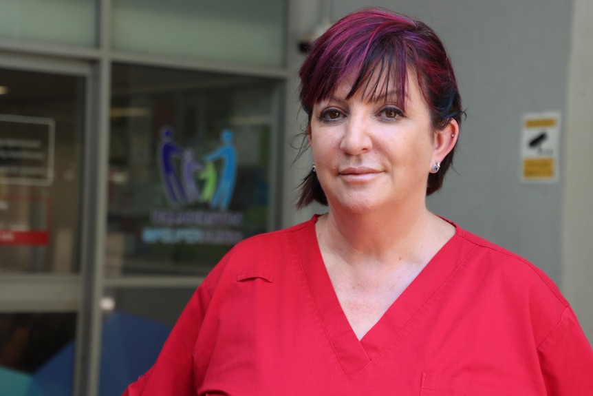 A serious-looking woman in red scrubs standing in front of a medical clinic building.