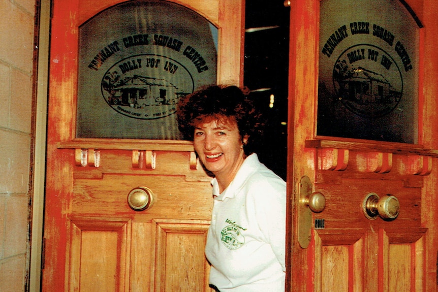 A woman in a white shirt smiles, peering out from between two wooden doors.