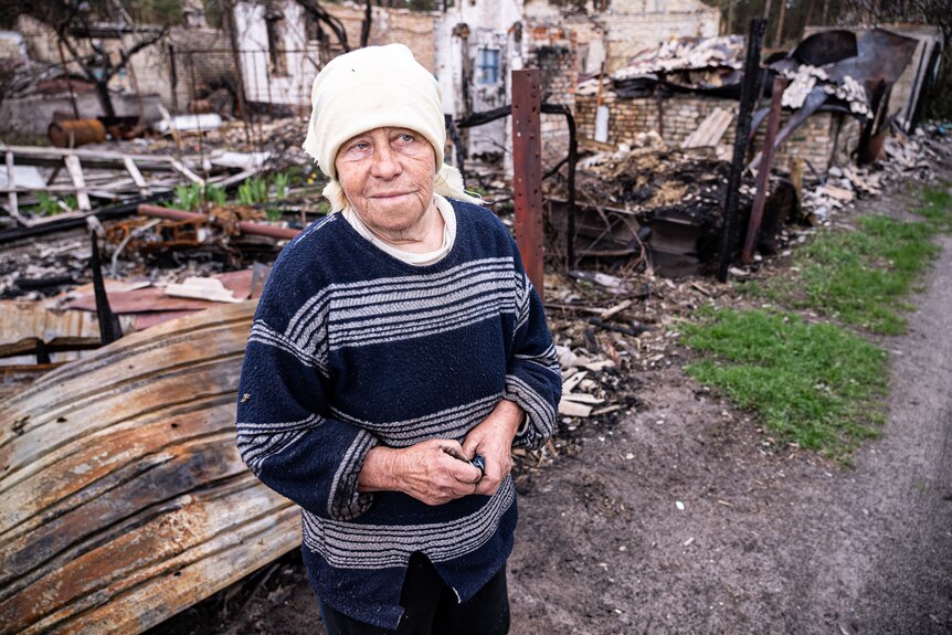 An older woman in a striped jumper and cream headscarf stands in front of burned out homes