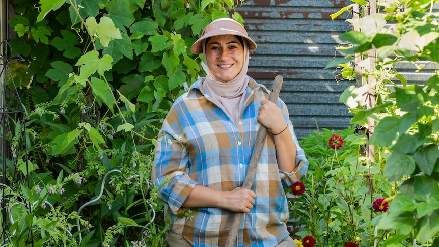 A woman wearing a hijab under a visor and holding a rake stands smiling proudly amongst a very green and lush garden