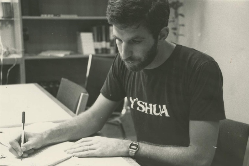 Black and white photograph of Bob Mendelsohn wearing Y'shua (Jesus) t-shirt and writing in notepad.