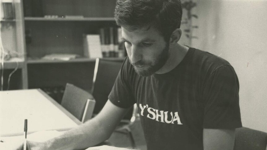 Black and white photograph of Bob Mendelsohn wearing Y'shua (Jesus) t-shirt and writing in notepad.